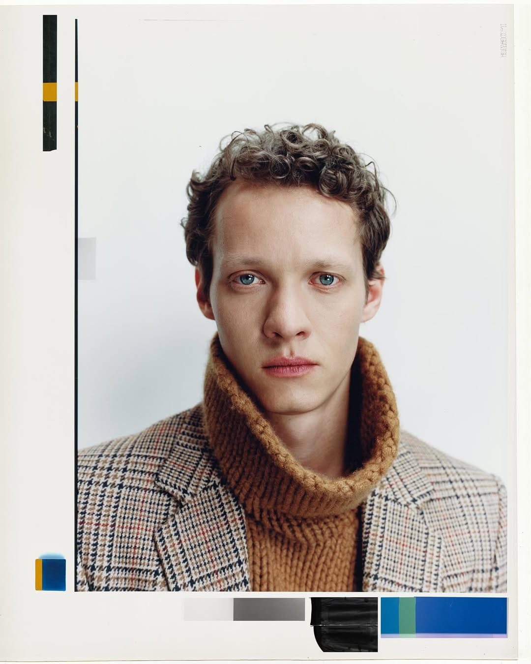 Portrait of a man with curly hair wearing a brown turtleneck sweater and patterned blazer against a white backdrop.