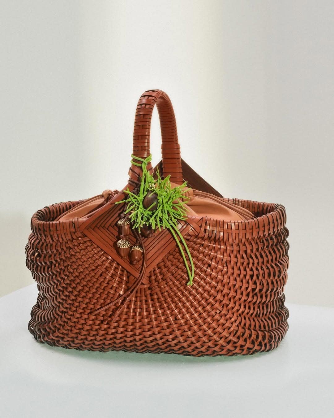 Brown woven leather basket handbag with acorn charms and bright green thread detail on a white background