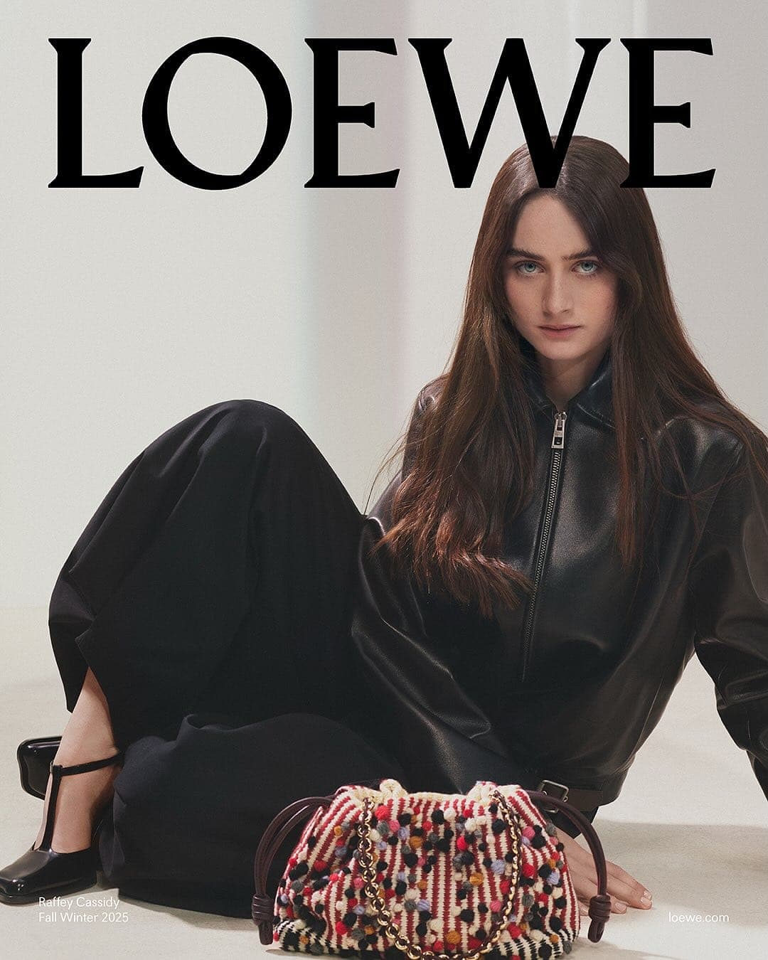 Model seated on the floor wearing a black leather jacket next to a colourful woven handbag for Loewe Fall 2025 ad campaign.