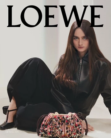 Model seated on the floor wearing a black leather jacket next to a colourful woven handbag for Loewe Fall 2025 ad campaign.