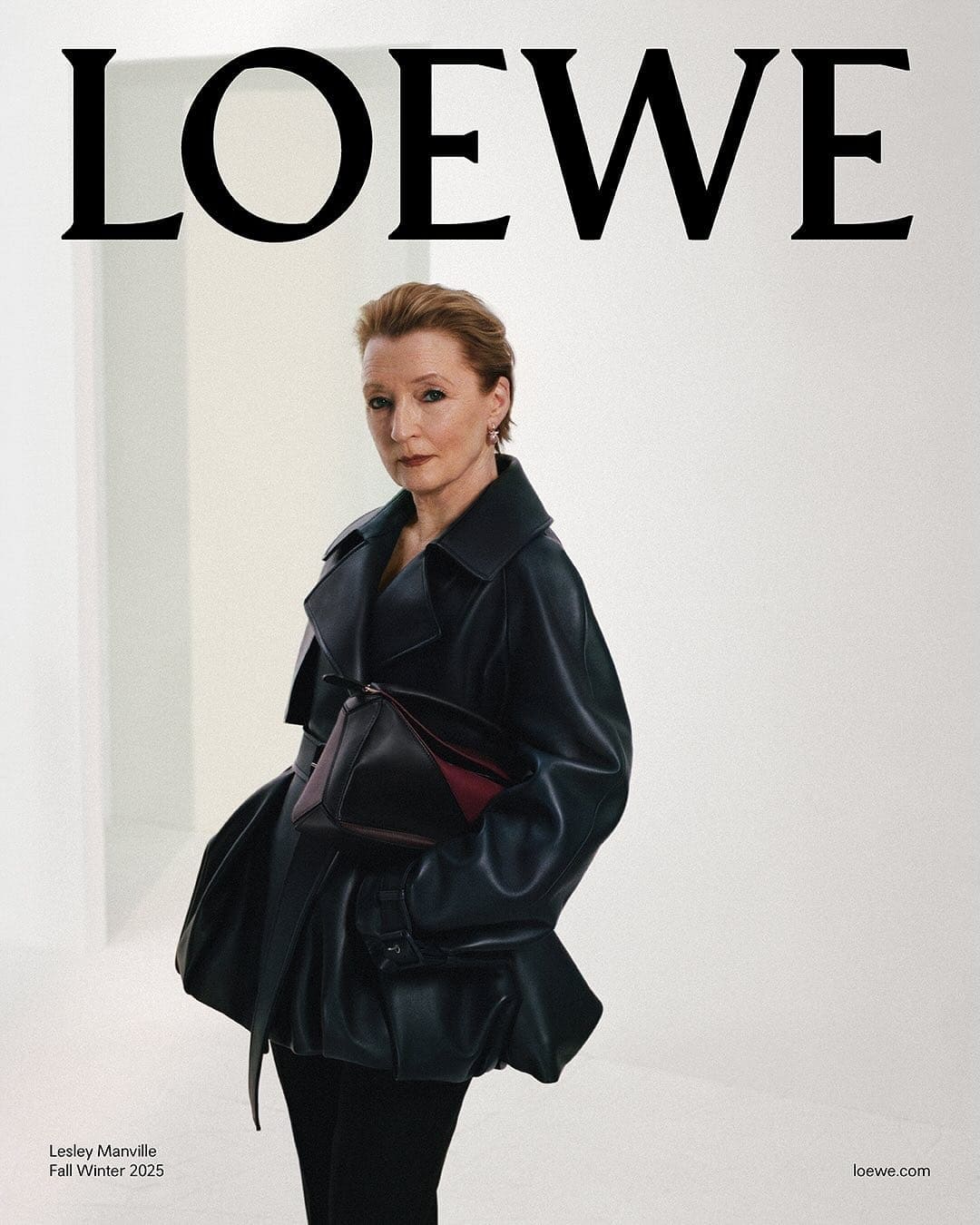 Lesley Manville wearing a black leather coat holding a dark handbag against a neutral white studio background