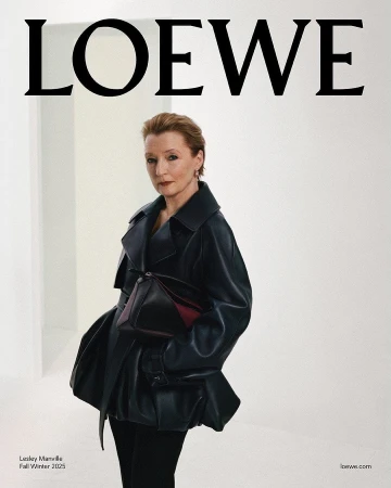 Lesley Manville wearing a black leather coat holding a dark handbag against a neutral white studio background