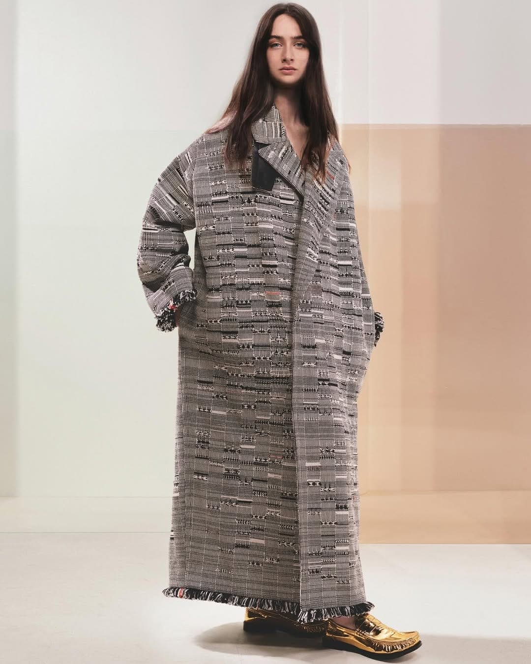 Model wearing a long patterned tweed coat and metallic gold shoes against a minimal studio background