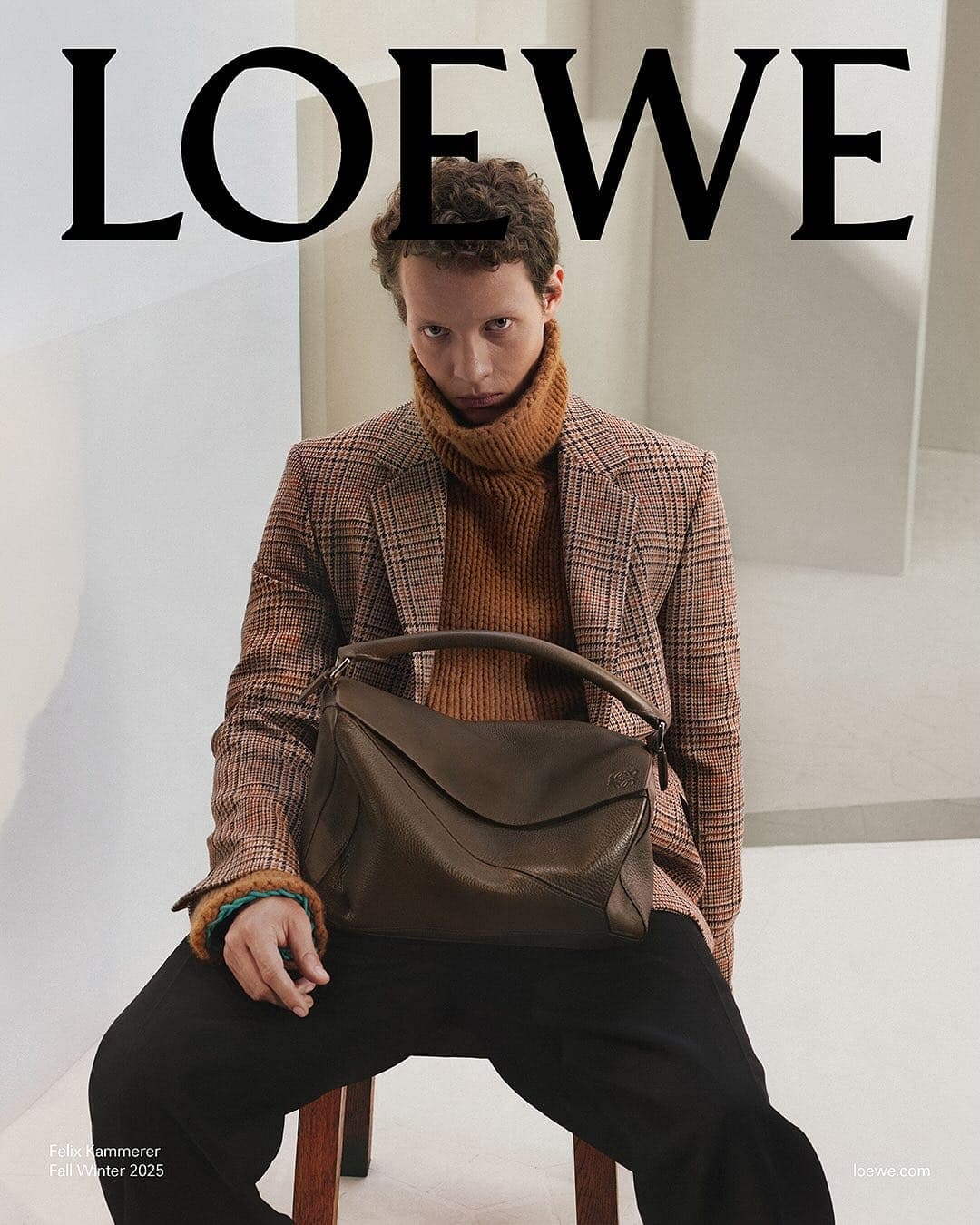 Man wearing a plaid blazer and brown turtleneck holding a dark leather handbag against an architectural studio background