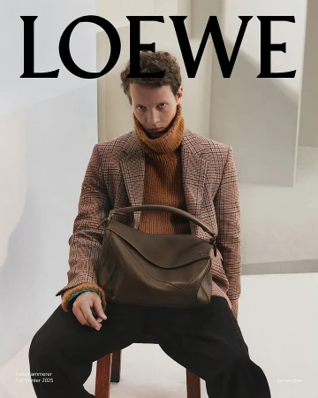 Man wearing a plaid blazer and brown turtleneck holding a dark leather handbag against an architectural studio background
