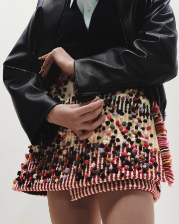 Model wearing a black leather jacket and a textured multi-colored fringed skirt against a white background