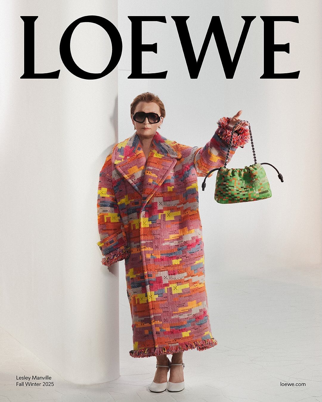 Lesley Manville wearing a vibrant patterned coat holding a green handbag in a white studio setting