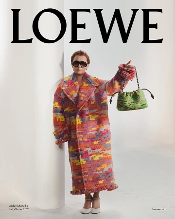 Lesley Manville wearing a vibrant patterned coat holding a green handbag in a white studio setting