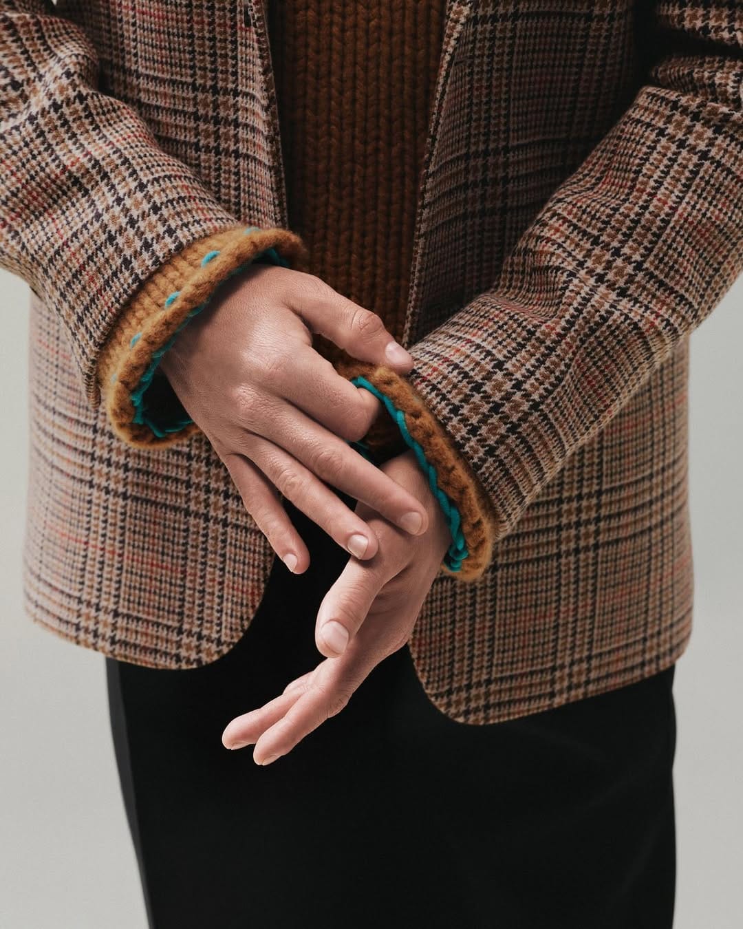 Close-up of hands adjusting a brown plaid blazer over a ribbed knit sweater against a plain grey studio background