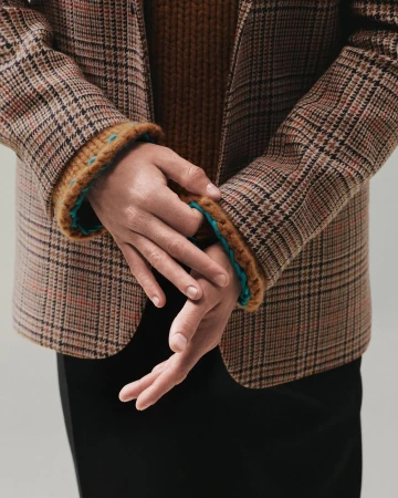 Close-up of hands adjusting a brown plaid blazer over a ribbed knit sweater against a plain grey studio background