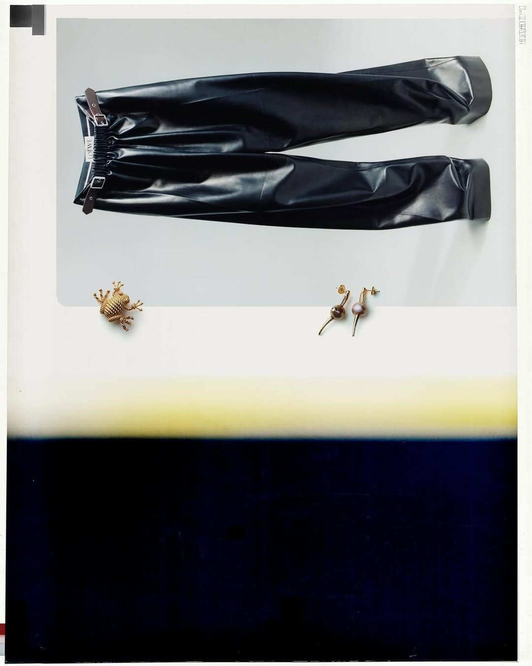 Loewe Fall 2025 ad campaign still showing dark leather trousers, a frog brooch, and earrings on a white and blue surface.
