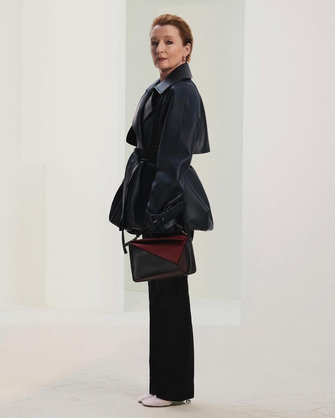 Lesley Manville wearing dark coat and holding a color-blocked handbag in a minimalist white architectural interior for Loewe Fall 2025 ad campaign