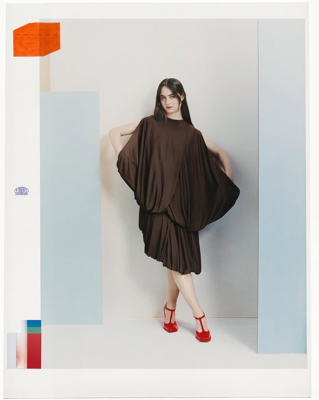 Model wearing a draped brown dress and red shoes standing against a geometric studio background for Loewe Fall 2025 campaign.