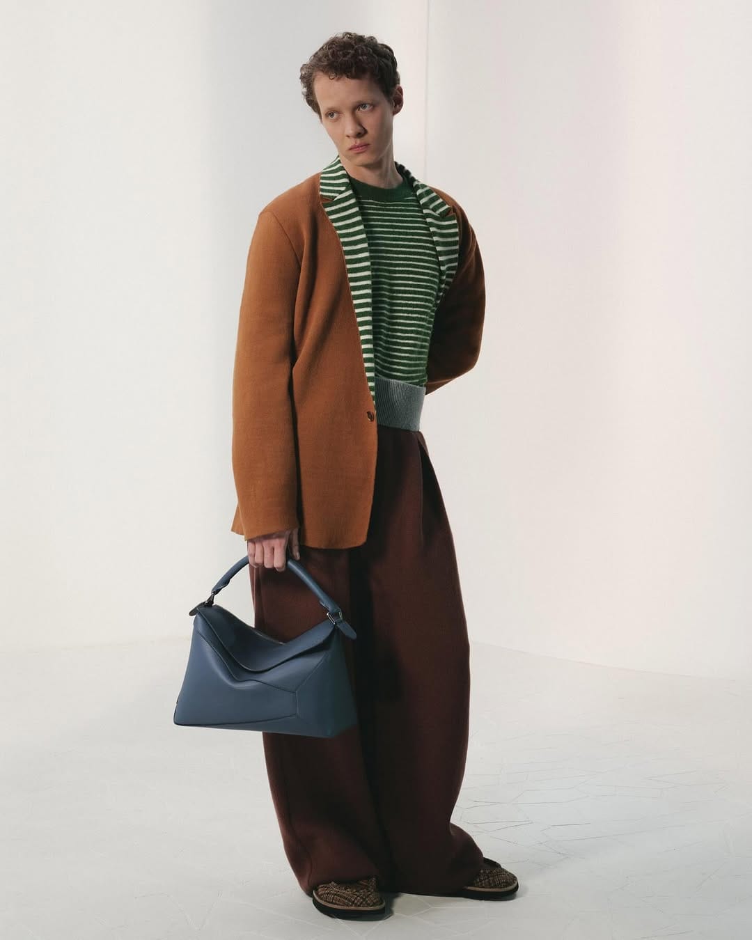Man wearing a brown blazer, striped sweater, and dark trousers holding a blue leather handbag against a plain white studio background.