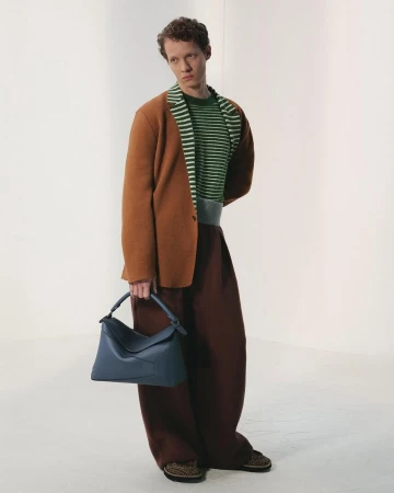 Man wearing a brown blazer, striped sweater, and dark trousers holding a blue leather handbag against a plain white studio background.
