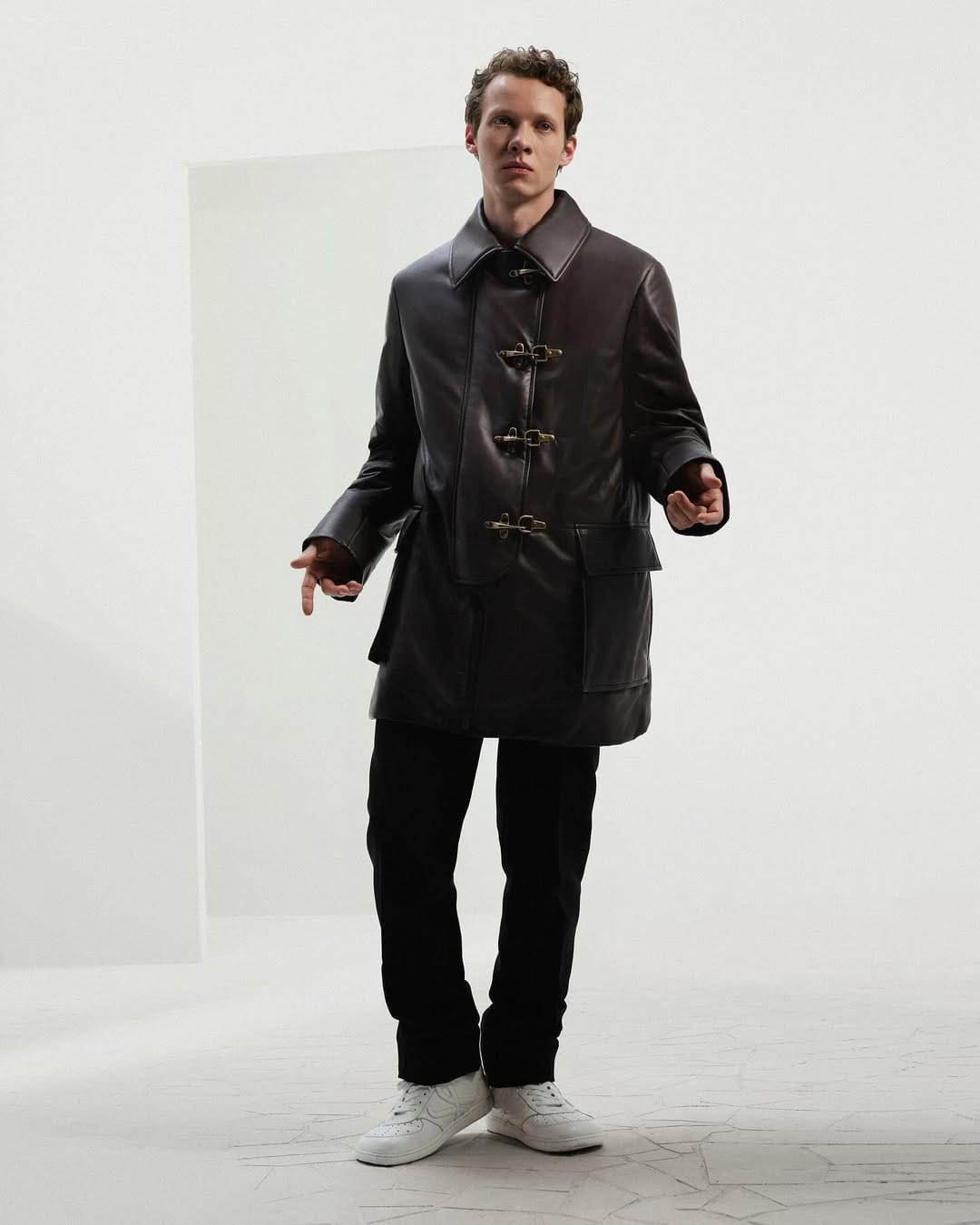A young man wearing a dark leather coat and white sneakers standing against a white seamless background