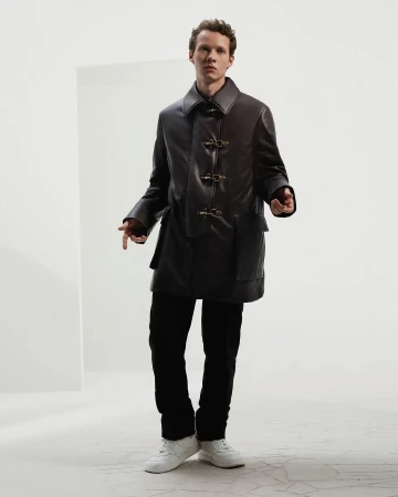 A young man wearing a dark leather coat and white sneakers standing against a white seamless background