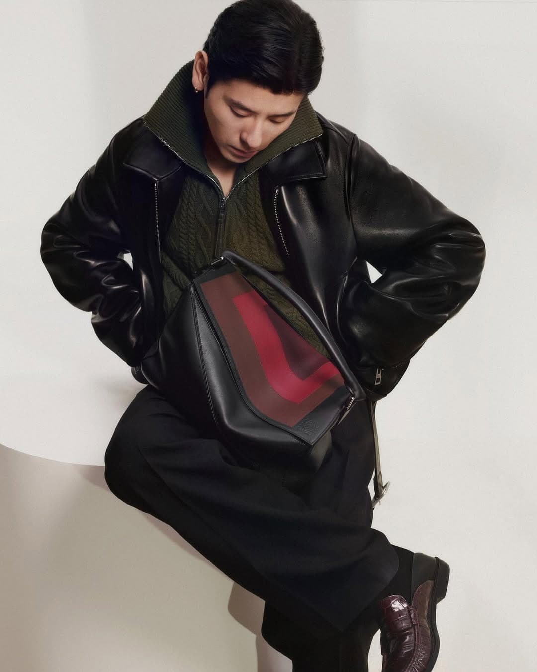Man wearing a black leather jacket and dark trousers holding a structured black leather handbag against a white background