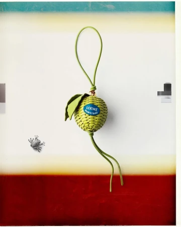 Woven green leather fruit charm featuring a blue brand label suspended against a white and red background.
