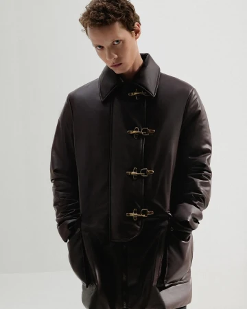 Model wearing a dark brown leather coat with brass toggle closures against a plain white studio background.