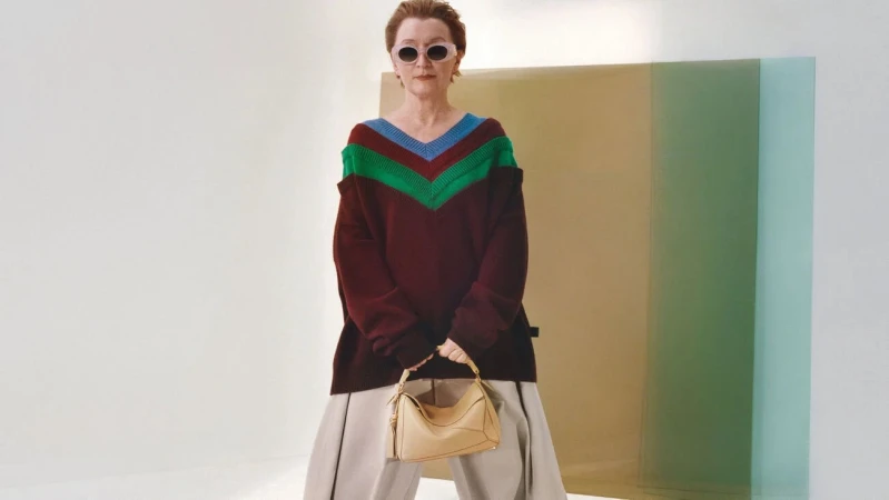 Elderly woman wearing sunglasses and a chevron knit sweater holding a light tan handbag against a white studio background