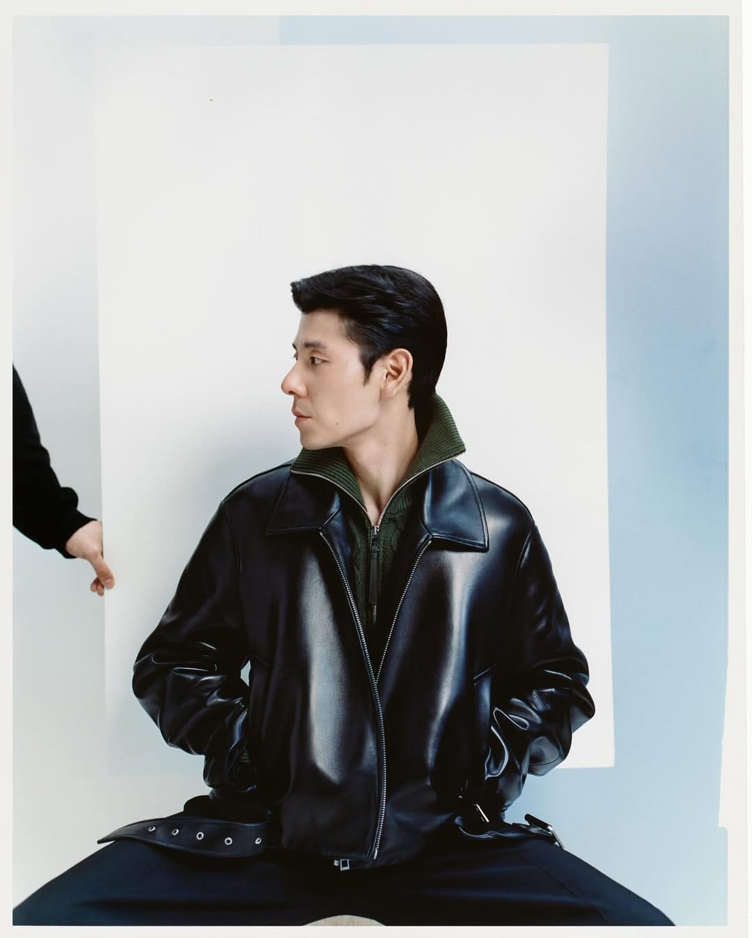 Man wearing a black leather jacket and olive green collar against a white and pale blue studio backdrop