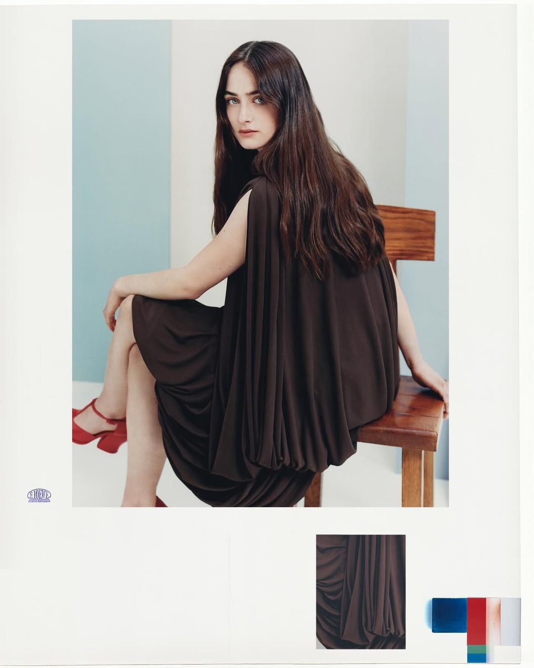 Woman with long dark hair wearing a dark brown draped dress and red platform shoes seated on a wooden chair.