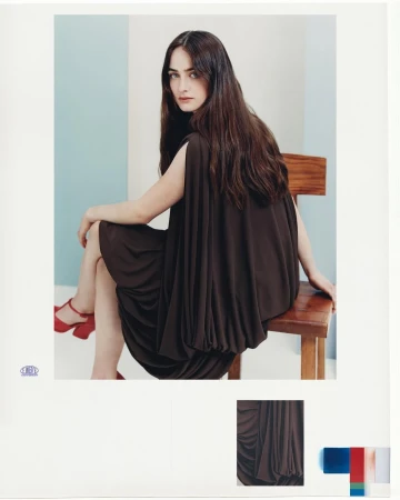 Woman with long dark hair wearing a dark brown draped dress and red platform shoes seated on a wooden chair.