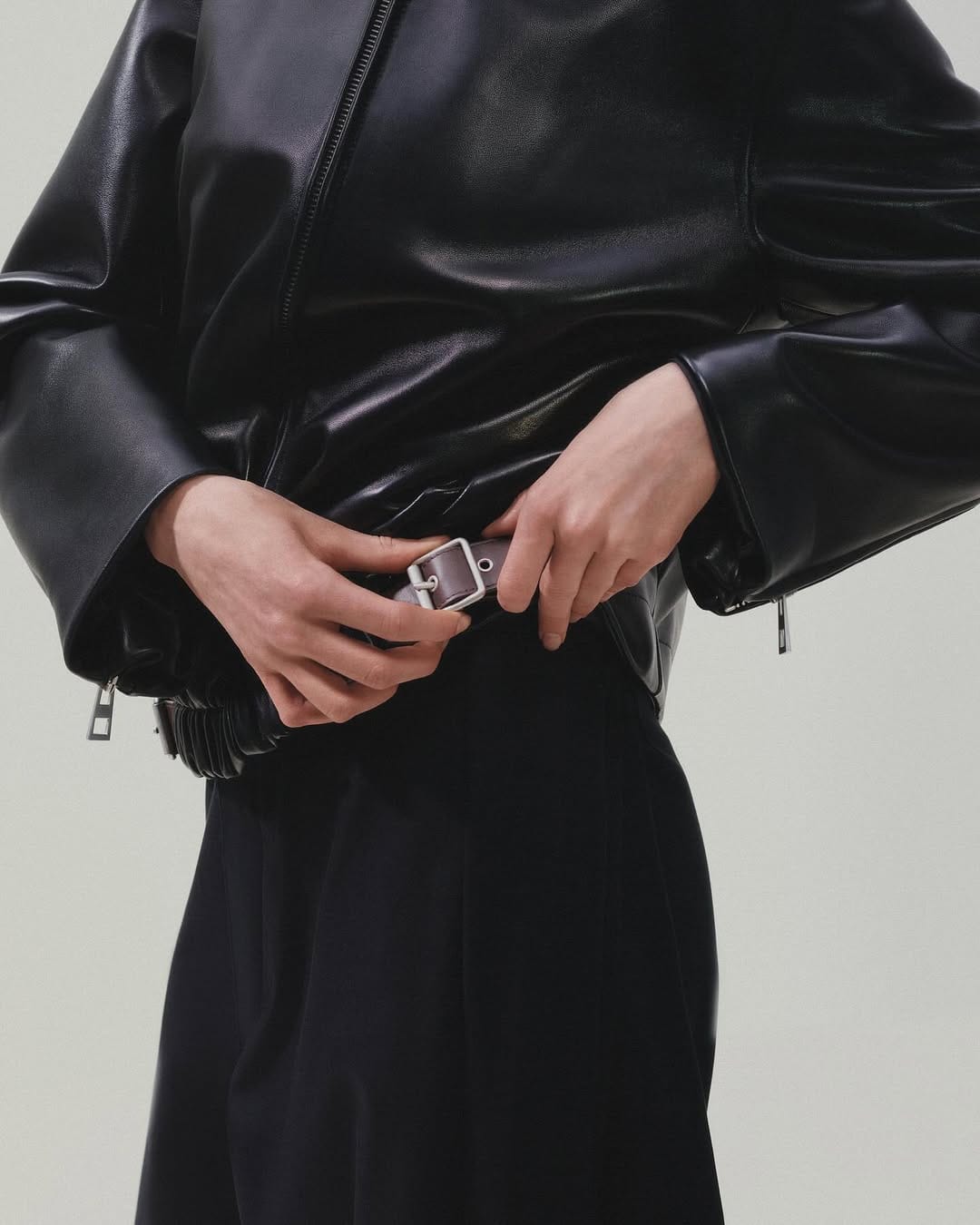 Close-up of a person wearing a black leather jacket and fastening a brown belt against a plain light background