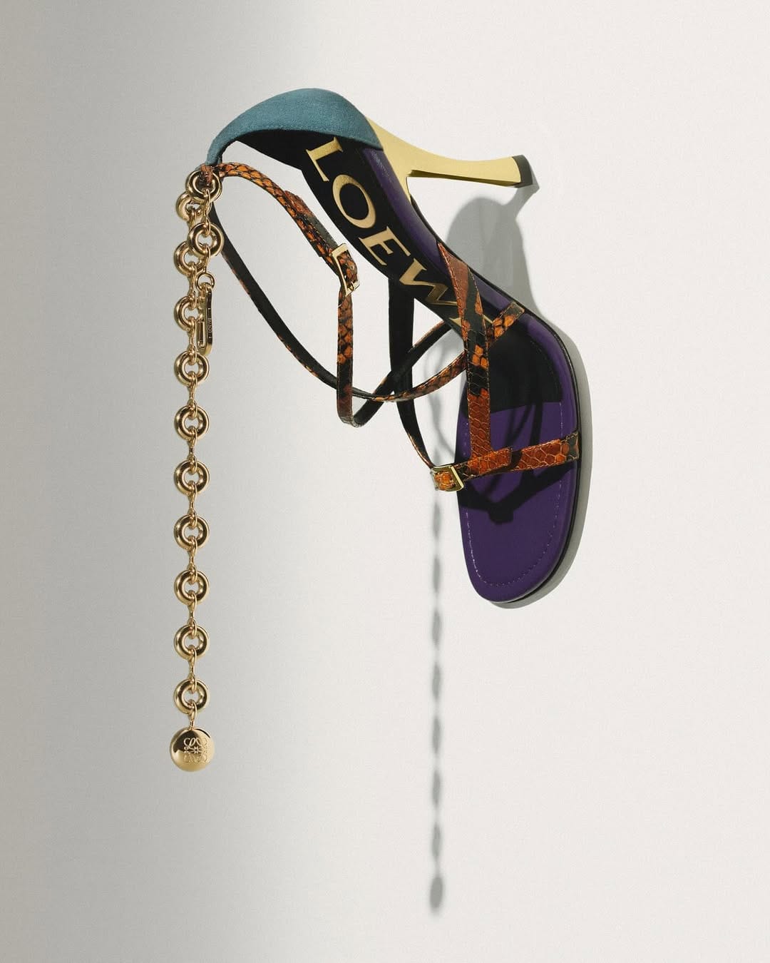 Loewe high-heeled sandal featuring purple sole, snakeskin straps, teal heel collar, and hanging gold chain with medallion against a white backdrop