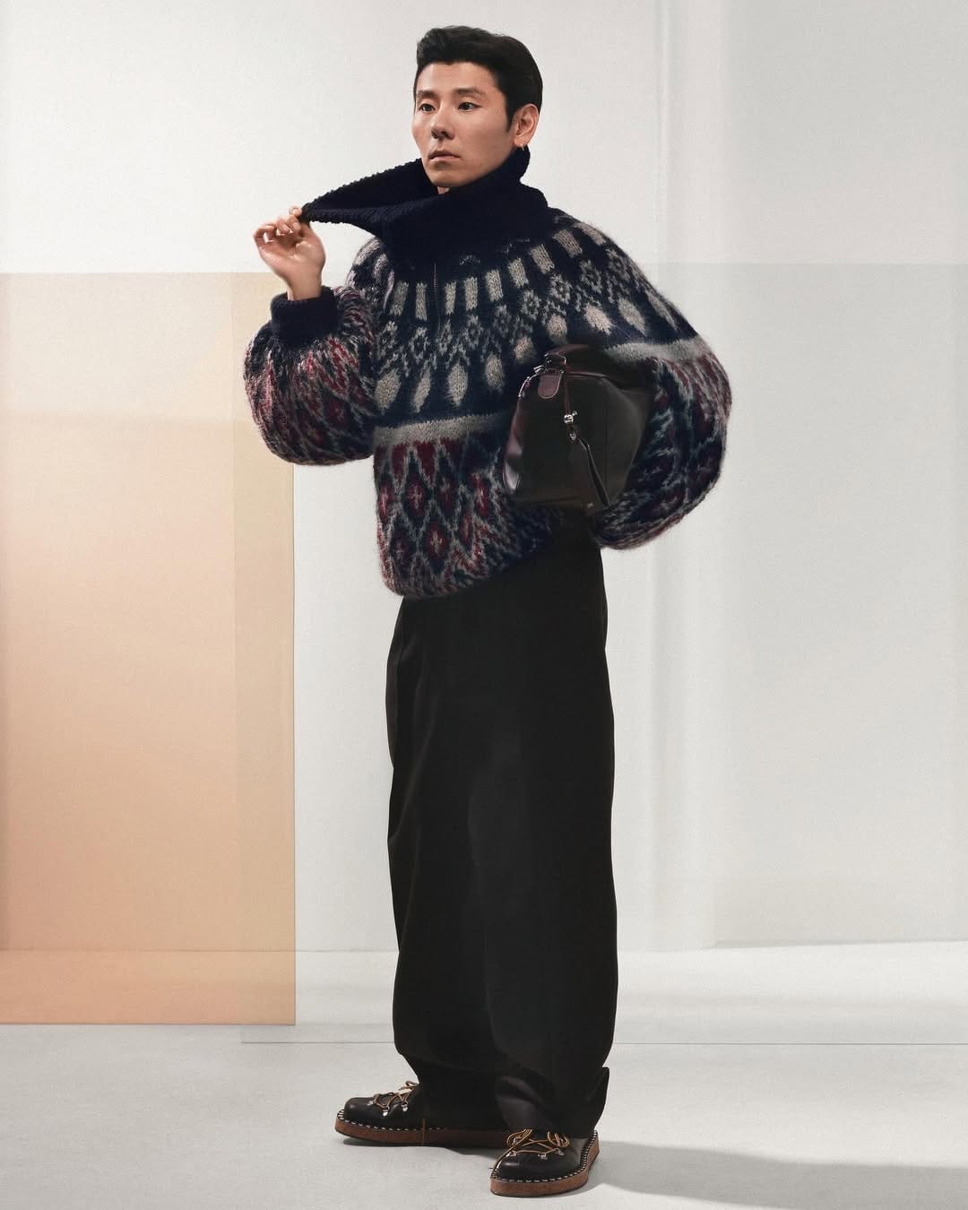 Man wearing a patterned sweater and dark trousers holding a dark handbag against a plain white studio backdrop.
