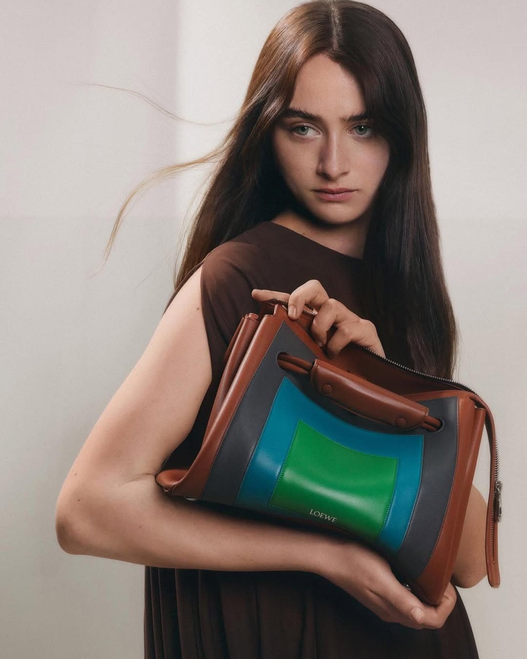 A woman holding a structured brown leather handbag with blue and green panels against a plain neutral background.