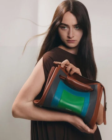A woman holding a structured brown leather handbag with blue and green panels against a plain neutral background.