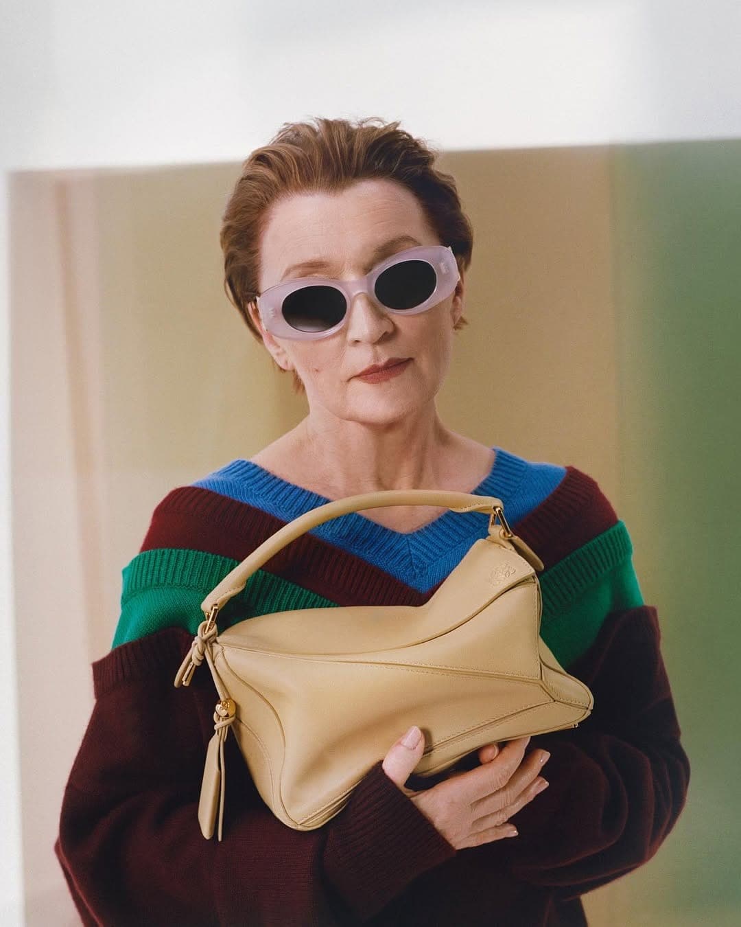 Older woman with short brown hair wearing translucent sunglasses and holding a light tan leather handbag against a color-blocked backdrop