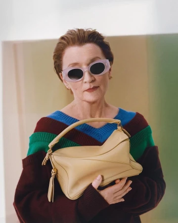 Older woman with short brown hair wearing translucent sunglasses and holding a light tan leather handbag against a color-blocked backdrop
