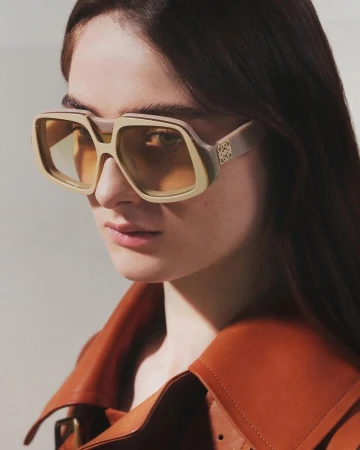 Close-up portrait of a woman wearing oversized beige sunglasses and a brown leather coat against a neutral background.