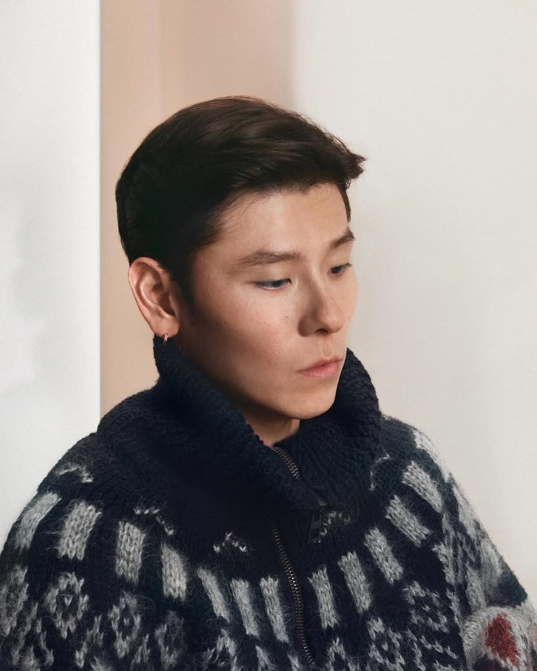 Close-up portrait of Jiang Qiming wearing a dark patterned knit turtleneck sweater against a neutral background.