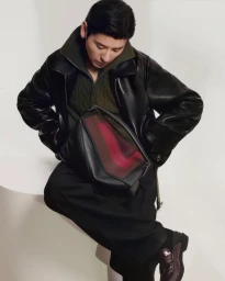 Man wearing a black leather jacket and dark trousers holding a structured black leather handbag against a white background