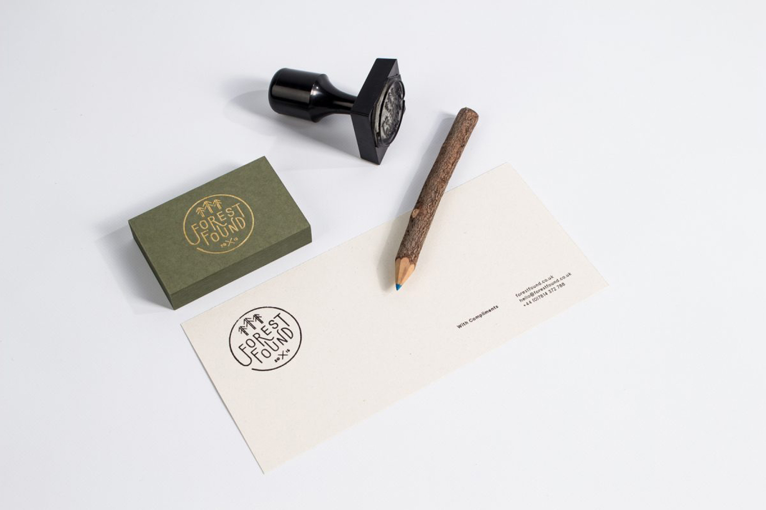 Branding mockup featuring an olive green business card, cream envelope, black stamp, and twig pencil on white background.