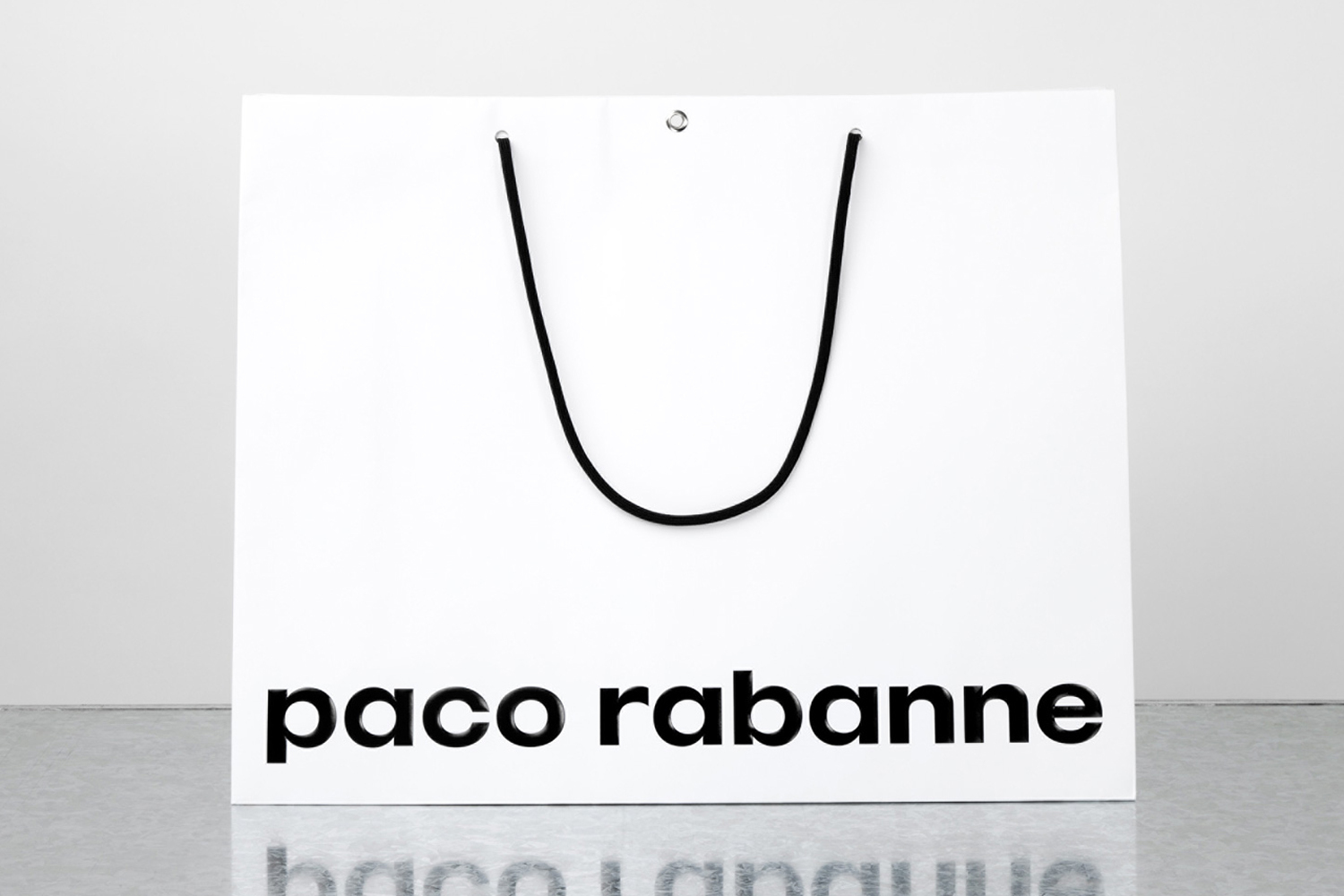 White shopping bag with black rope handles and Paco Rabanne wordmark on a reflective grey surface against a seamless background.