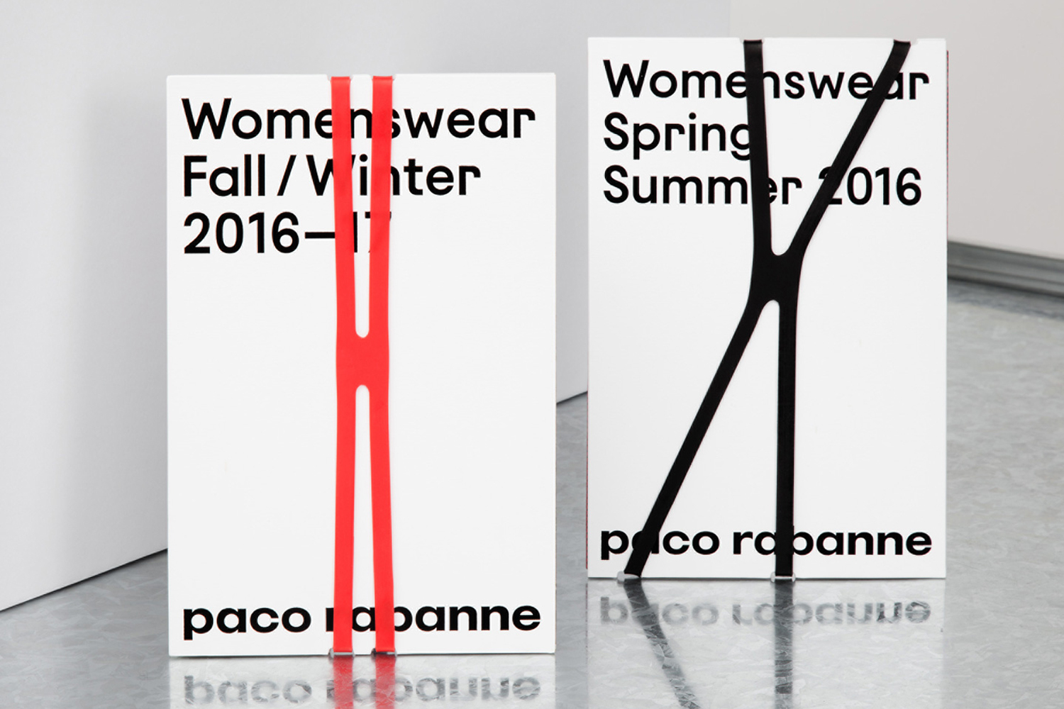 Two Paco Rabanne lookbooks featuring white covers with bold black typography, bound with red and black elastic bands on a reflective surface.