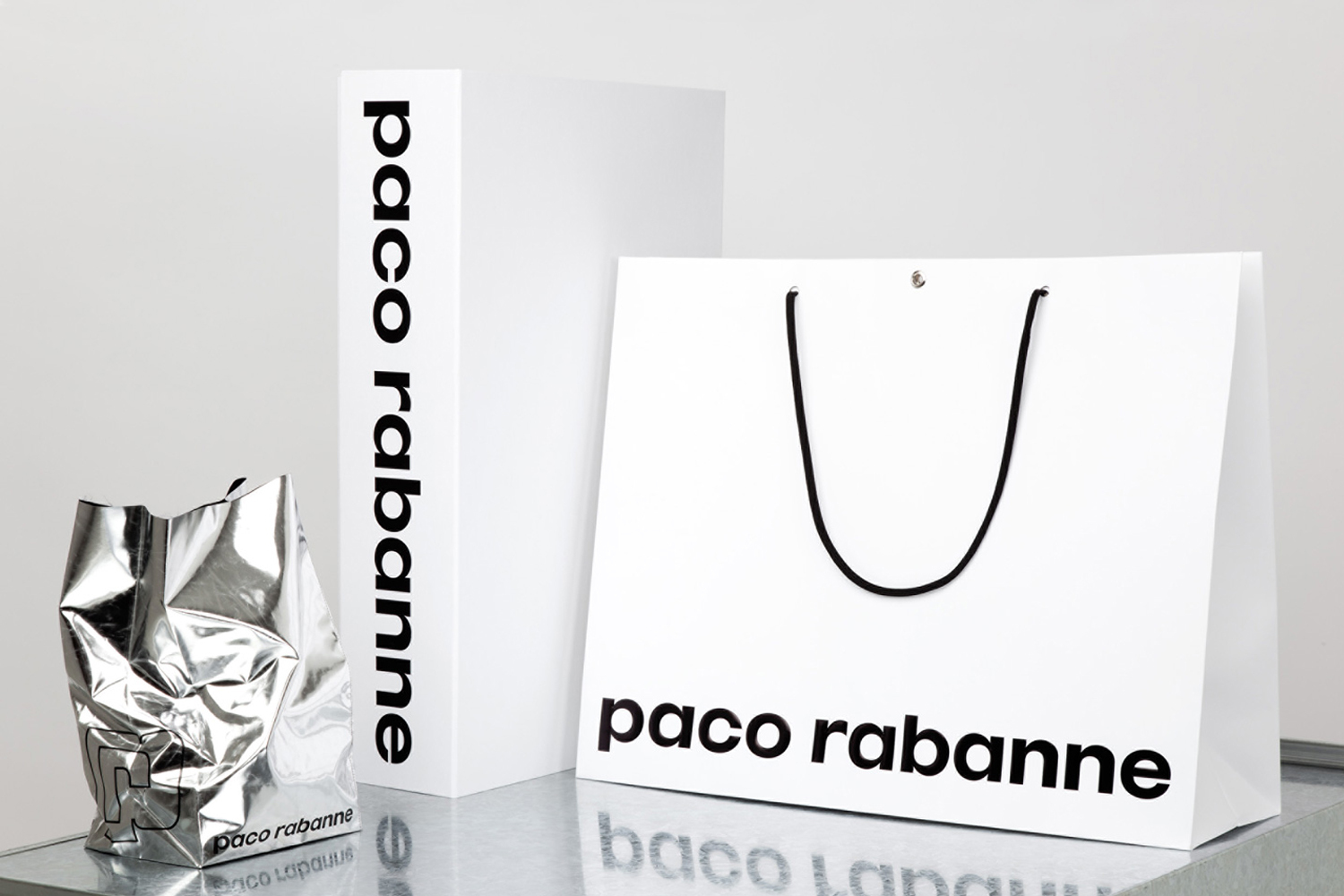 Paco Rabanne brand identity retail packaging including white shopping bags and a metallic silver bag on a reflective surface