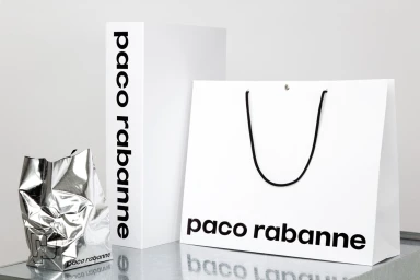 Paco Rabanne brand identity retail packaging including white shopping bags and a metallic silver bag on a reflective surface
