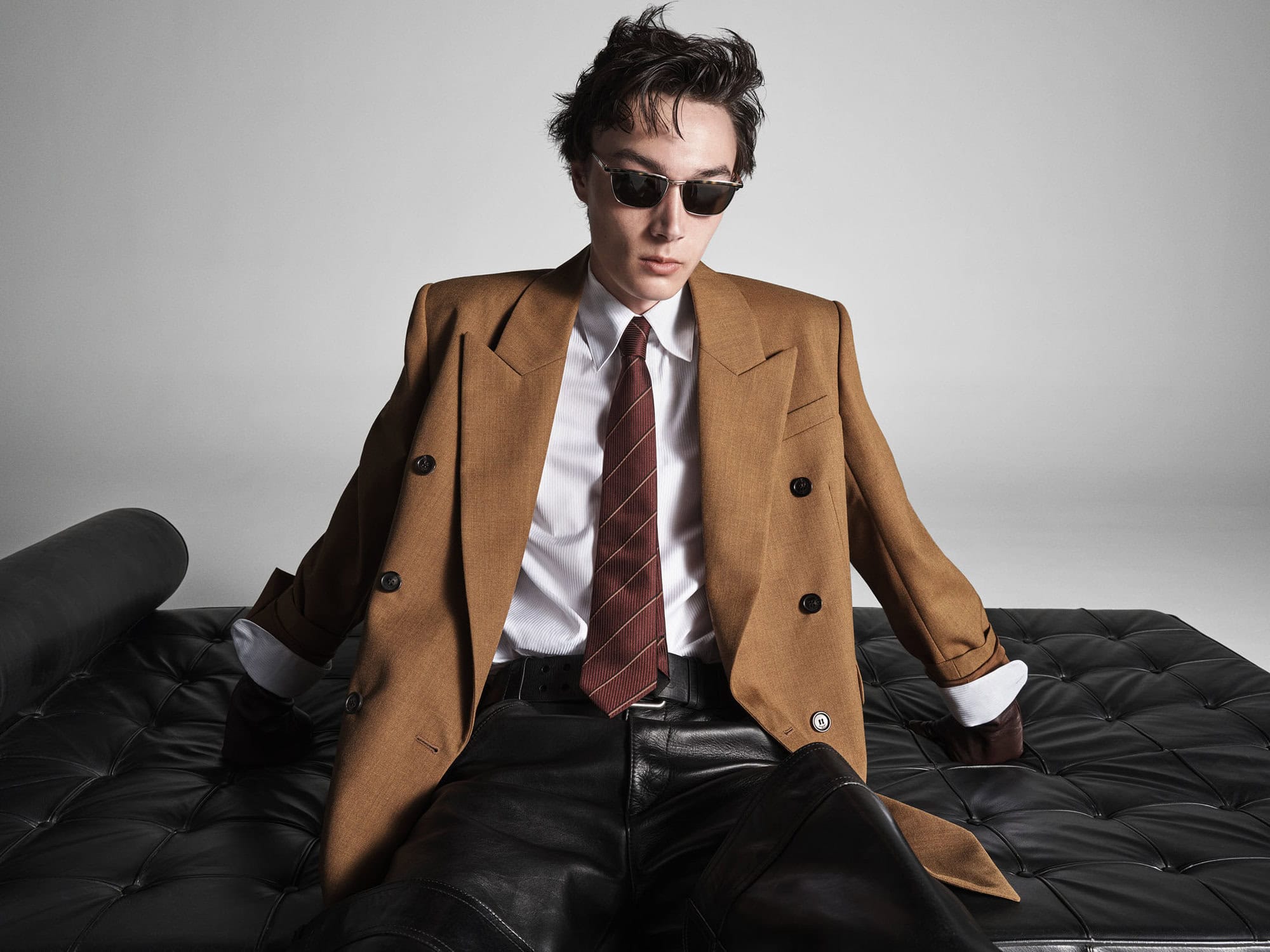 Man wearing sunglasses, brown blazer, white shirt, and tie sitting on black tufted leather furniture.
