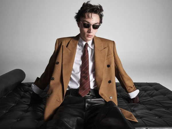 Man wearing sunglasses, brown blazer, white shirt, and tie sitting on black tufted leather furniture.