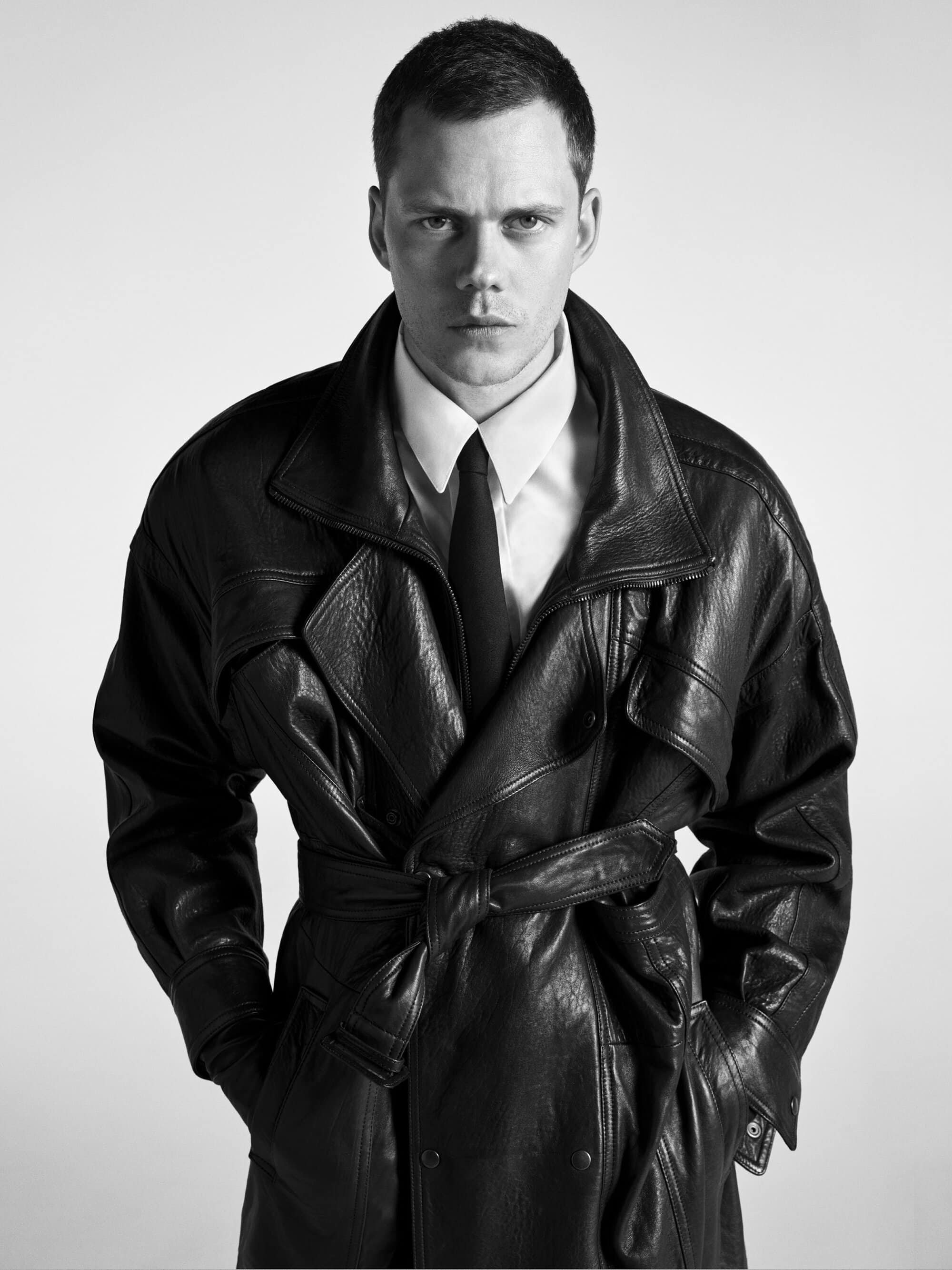 Bill Skarsgard wearing a black leather trench coat, white shirt, and tie against a plain light background