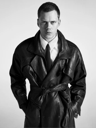 Bill Skarsgard wearing a black leather trench coat, white shirt, and tie against a plain light background