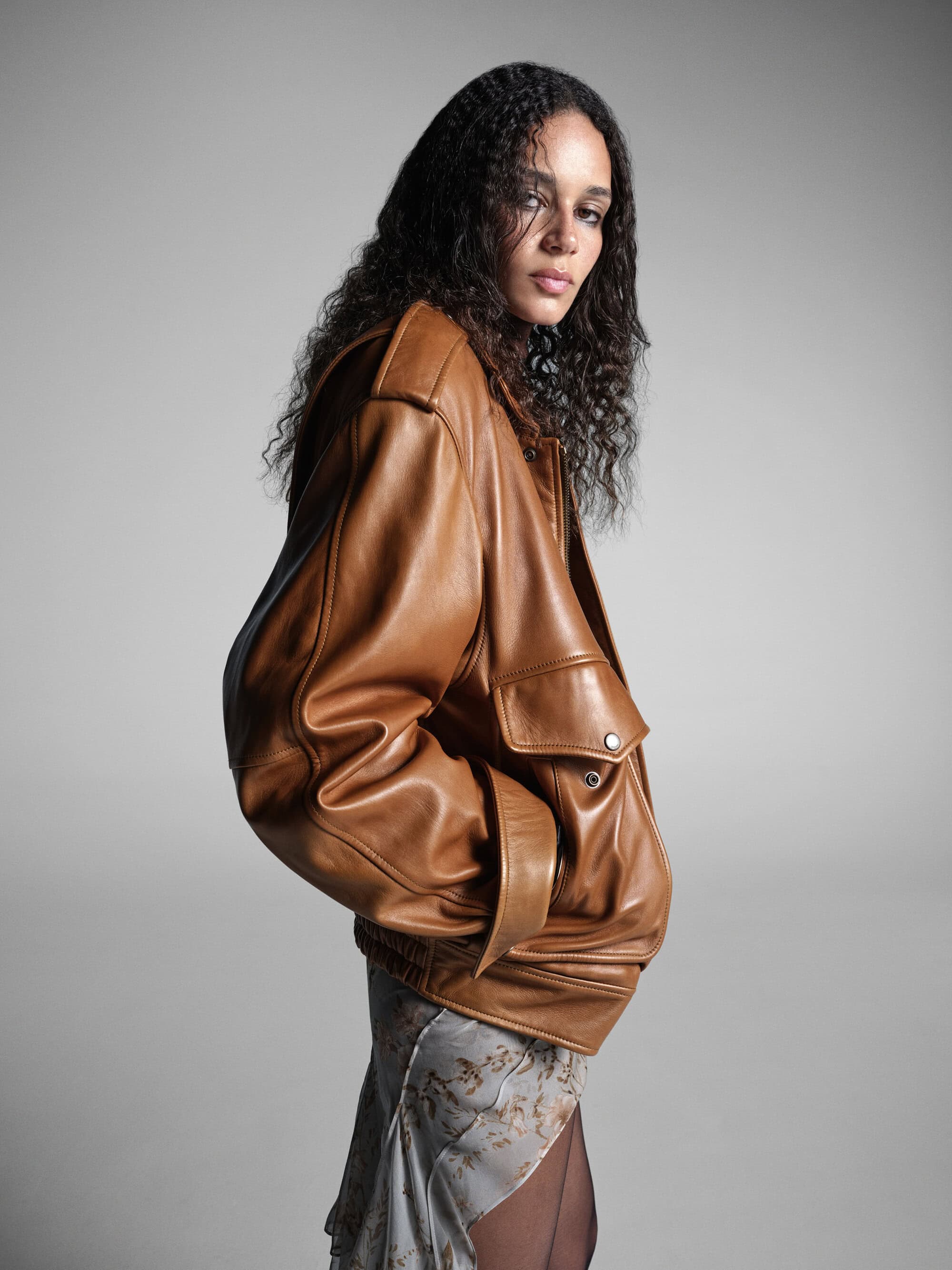 Model with long curly hair wearing an oversized brown leather jacket against a neutral grey studio background
