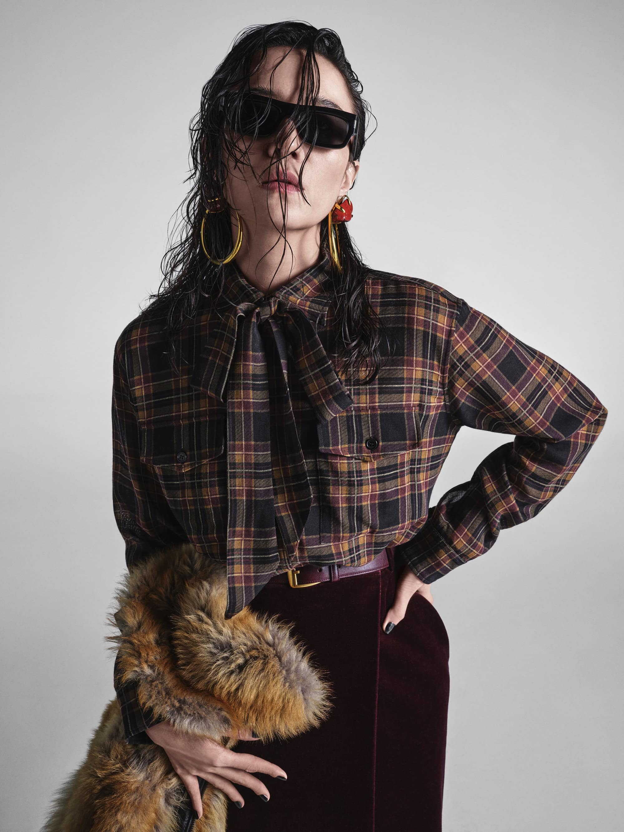 Model with wet hair wearing sunglasses and a plaid blouse holding a fur piece against a grey background.