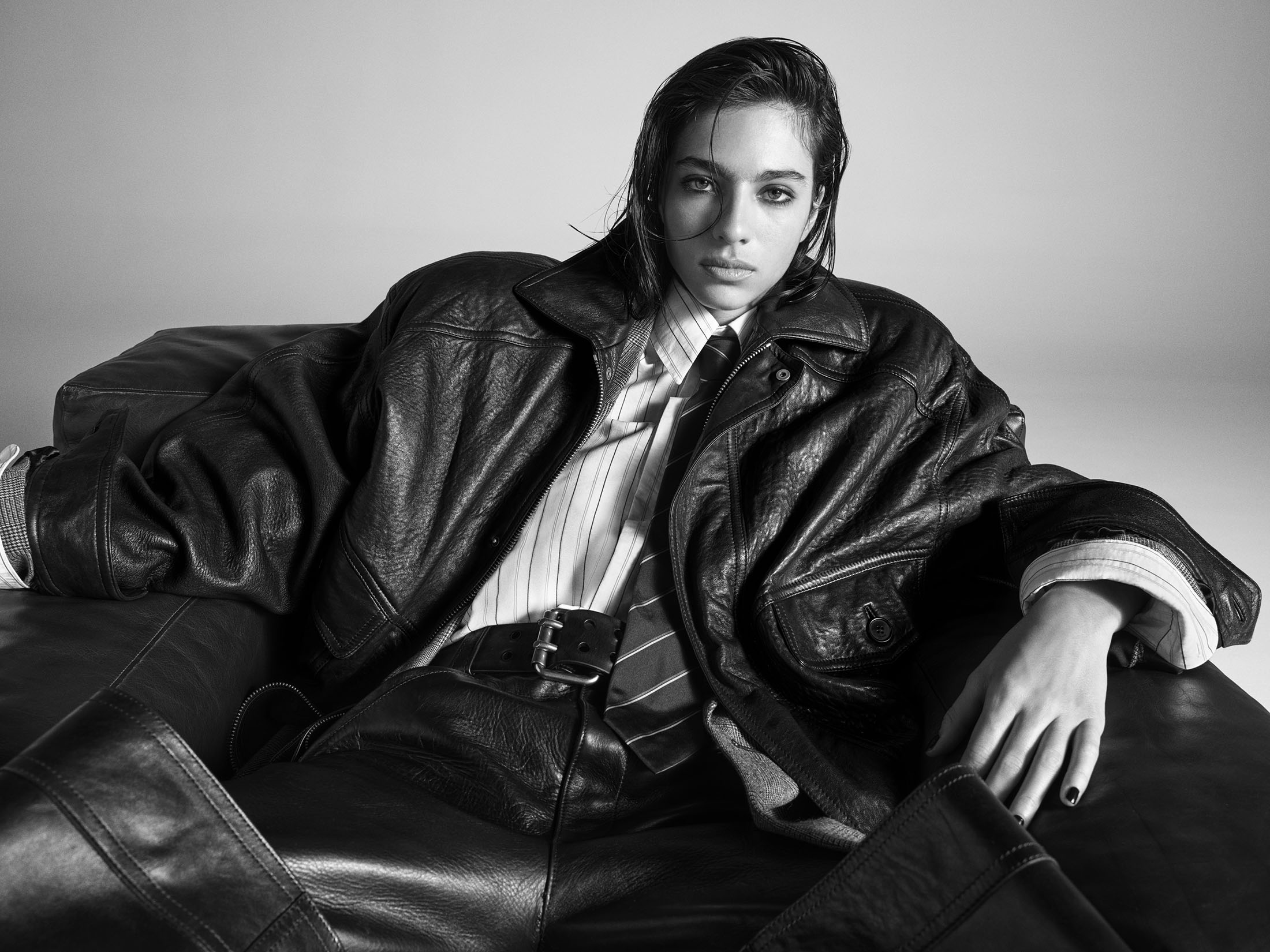 Model seated on a dark leather couch wearing an oversized leather jacket, collared shirt, and tie against a plain studio background.